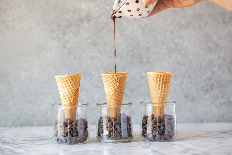 drizzling melted chocolate into the bottom of 3 sugar ice cream cones standing in glass jars filled with chocolate chips