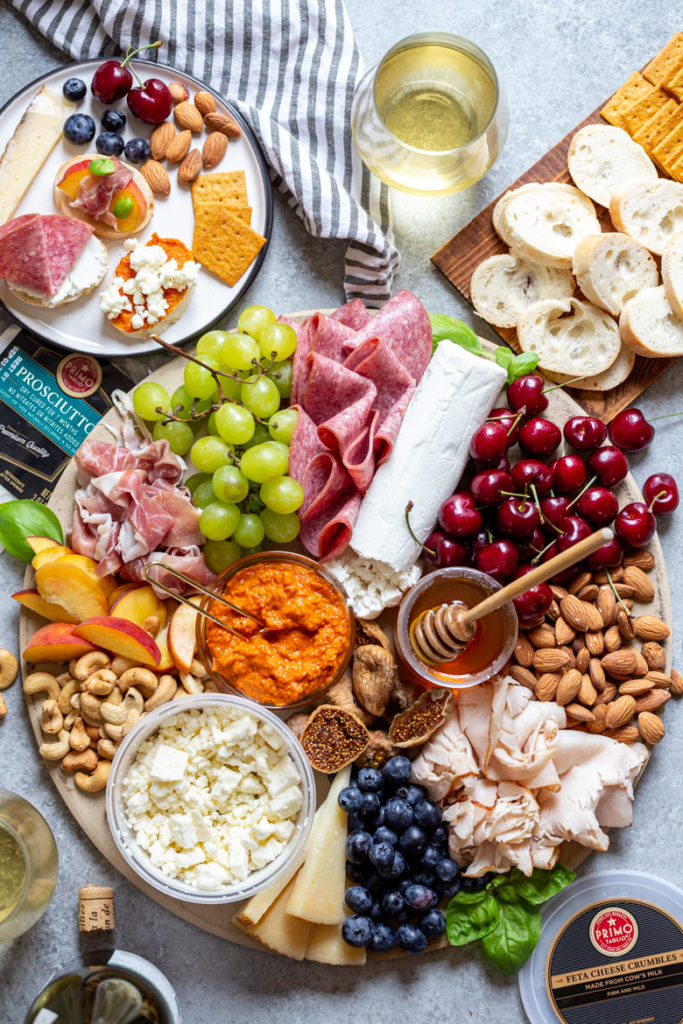 Summer produce with meats and cheeses styled onto a round cutting board on a concrete surface