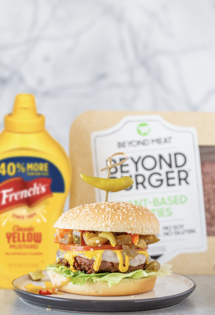 Beyond Burger plant-based patty piled high with provolone cheese, mustard, lettuce, tomato and giardiniera all on a seeded bun with a package of Beyond Meat Patties and French's yellow mustard