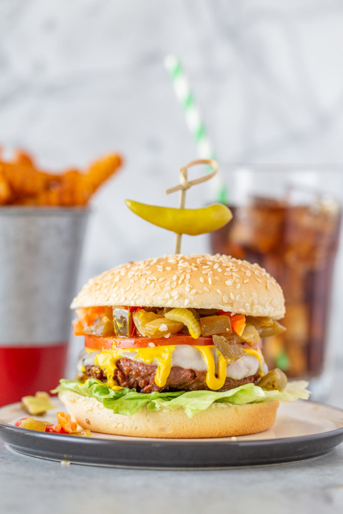 These Chicago-Style Beyond Burgers are a plant-based patty piled high with provolone cheese, mustard, lettuce, tomato and giardiniera all on a seeded bun with a container of french fries and a coke with a paper straw