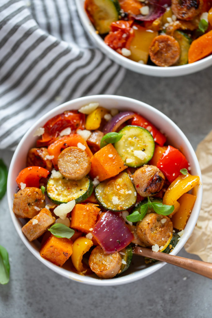 Bowl filled with oven roasted vegetables and chicken sausages, topped with fresh basil and parmesan cheese